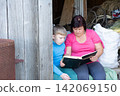 Woman and boy sharing a moment reading a book together in the countryside, seated by a wooden barn door. Nostalgic scene. 142069150