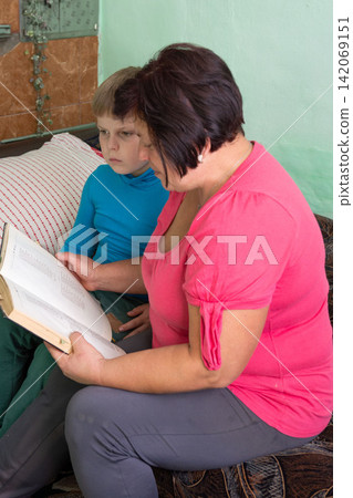 Grandmother and grandson share a moment reading together indoors, fostering learning and connection in a cozy setting. Grandmother and grandson share a moment reading together indoors, fostering learning and connection in a cozy setting. 142069151