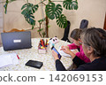 A woman assists a child with homework at a table with a laptop, plant, and art supplies. The woman uses her phone to help the child. 142069153