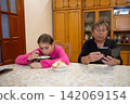 Grandmother and granddaughter at the dinner table, the girl is eating soup while the grandmother is on her phone 142069154