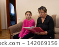 Grandmother and granddaughter share a book on a neutral-toned sofa, creating a heartwarming intergenerational bonding experience. 142069156