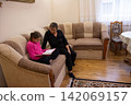 Grandmother reading a book to her granddaughter sitting on the couch in the living room, creating a heartwarming intergenerational connection. 142069157