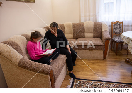 Grandmother reading a book to her granddaughter sitting on the couch in the living room, creating a heartwarming intergenerational connection. Grandmother reading a book to her granddaughter sitting on the couch in the living room, creating a heartwarming intergenerational connection. 142069157