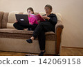 Grandmother and granddaughter working on laptop and reading a binder together while sitting on a couch in the living room. 142069158