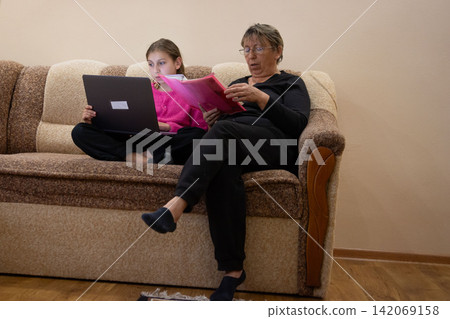Grandmother and granddaughter working on laptop and reading a binder together while sitting on a couch in the living room. Grandmother and granddaughter working on laptop and reading a binder together while sitting on a couch in the living room. 142069158