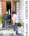 A boy with a backpack is leaving his house while a woman watches. Both are on the front steps of a home. He is on his way to school. 142069196