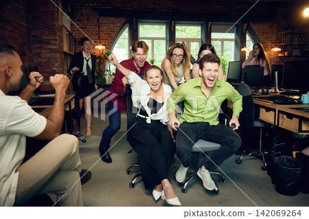Excited coworkers racing office chairs while colleagues cheer in modern workspace Excited coworkers racing office chairs while colleagues cheer in modern workspace 142069264