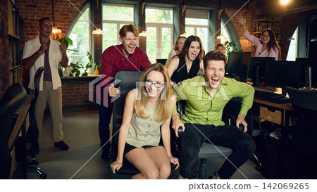 Cheerful coworkers celebrating during playful office chair race in creative workspace Cheerful coworkers celebrating during playful office chair race in creative workspace 142069265