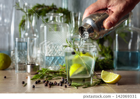 Gin and tonic cocktail is poured from a shaker into a glass with ice and lime wedges. Gin and tonic cocktail is poured from a shaker into a glass with ice and lime wedges. 142069331