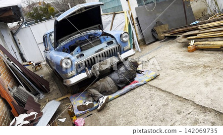 Male repairman lies under his vintage car at courtyard of house. Auto mechanic repairing his blue retro vehicle outdoor. Concept of automobile maintenance. Slow motion Male repairman lies under his vintage car at courtyard of house. Auto mechanic repairing his blue retro vehicle outdoor. Concept of automobile maintenance. Slow motion 142069735