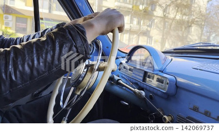 Male hands holds steering wheel while driving retro vehicle at urban street. Man riding on blue vintage auto at city road on sunny day. Driver moving in his old auto on route. Slow motion Male hands holds steering wheel while driving retro vehicle at urban street. Man riding on blue vintage auto at city road on sunny day. Driver moving in his old auto on route. Slow motion 142069737