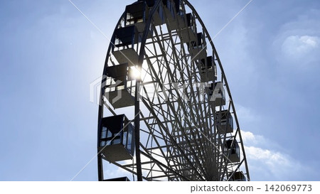 View of a ferris wheel against a clear blue sky with sunlight at background. The sun rays pass through the structure, creating bright highlights and a vibrant summer atmosphere around the attraction View of a ferris wheel against a clear blue sky with sunlight at background. The sun rays pass through the structure, creating bright highlights and a vibrant summer atmosphere around the attraction 142069773
