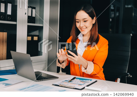 Smiling asian businesswoman talking on phone and using laptop while working at office desk Smiling asian businesswoman talking on phone and using laptop while working at office desk 142069940