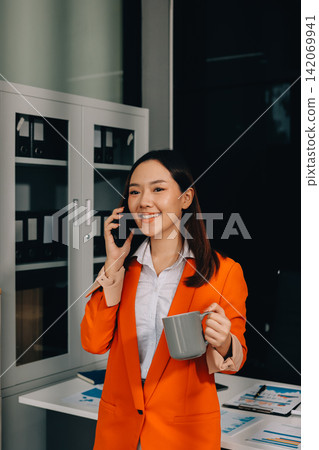 Smiling asian businesswoman talking on phone and using laptop while working at office desk Smiling asian businesswoman talking on phone and using laptop while working at office desk 142069941