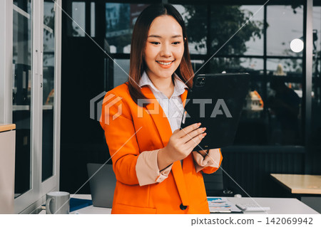 Smiling asian businesswoman talking on phone and using laptop while working at office desk Smiling asian businesswoman talking on phone and using laptop while working at office desk 142069942