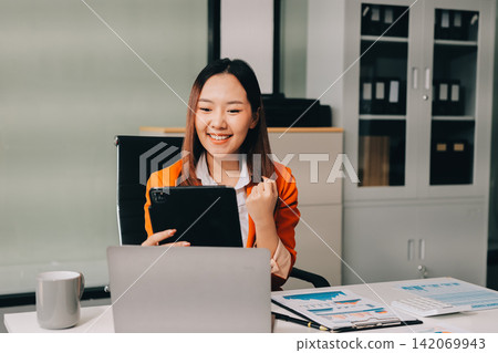 Smiling asian businesswoman talking on phone and using laptop while working at office desk 142069943