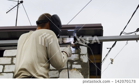 Manual laborer in protective mask welds iron detail at construction. Professional young repairman with welding machine working at the site. Male welder operating with metal on house. Close up Manual laborer in protective mask welds iron detail at construction. Professional young repairman with welding machine working at the site. Male welder operating with metal on house. Close up 142070178