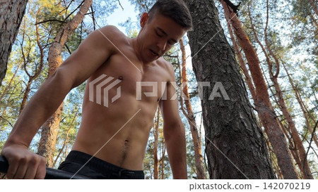 Hardy shirtless sportsman doing push ups on horizontal bar at summer forest. Strong and muscular guy training at nature. Young athletic man exercising outdoor. Concept of active and healthy lifestyle Hardy shirtless sportsman doing push ups on horizontal bar at summer forest. Strong and muscular guy training at nature. Young athletic man exercising outdoor. Concept of active and healthy lifestyle 142070219