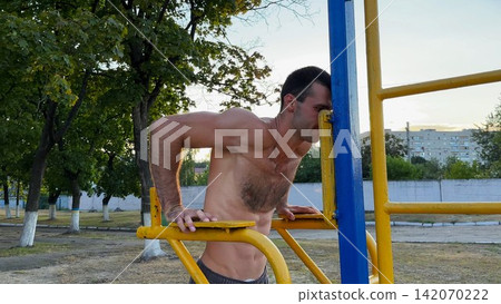 Strong sporty man exercising at sports ground. Male shirtless athlete doing push ups on parallel bars at sunny day. Young sportsman training hard outdoor. Concept of active sportive lifestyle Strong sporty man exercising at sports ground. Male shirtless athlete doing push ups on parallel bars at sunny day. Young sportsman training hard outdoor. Concept of active sportive lifestyle 142070222