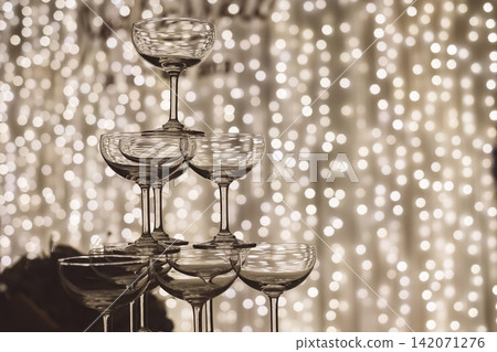 a silhouette shot of small tower of champagne's glass in wedding reception party near by a big cake and golden light bulbs in background. a silhouette shot of small tower of champagne's glass in wedding reception party near by a big cake and golden light bulbs in background. 142071276