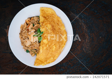 homemade Thai style spicy fried rice with stir-fried chicken and basil cover by a half circle omelet in white plate with spoon in white napkin on wood Serving trays with dark gray background homemade Thai style spicy fried rice with stir-fried chicken and basil cover by a half circle omelet in white plate with spoon in white napkin on wood Serving trays with dark gray background 142071571