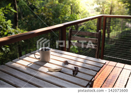 a coffee cup and many dried leaves on wooden table and chair at balcony with green trees in background in afternoon a coffee cup and many dried leaves on wooden table and chair at balcony with green trees in background in afternoon 142071600