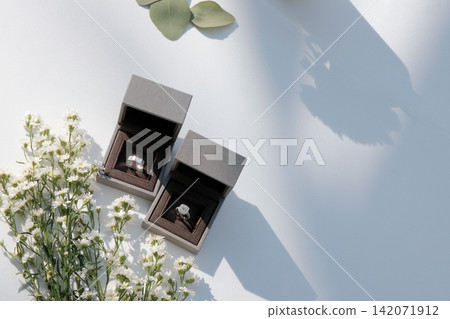 The wedding rings are in boxes on a corner of a white table, decorated with dried flowers, basking in the morning sunlight and shadow in a blank space. The wedding rings are in boxes on a corner of a white table, decorated with dried flowers, basking in the morning sunlight and shadow in a blank space. 142071912