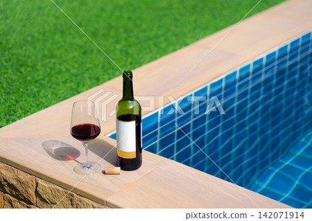 A bottle of red wine and a glass of wine are set on the edge of a clear blue pool, surrounded by a lush green lawn. The afternoon sun shines down, creating a relaxing and inviting atmosphere. A bottle of red wine and a glass of wine are set on the edge of a clear blue pool, surrounded by a lush green lawn. The afternoon sun shines down, creating a relaxing and inviting atmosphere. 142071914