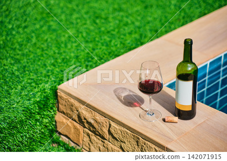 A bottle of red wine and a glass of wine are set on the edge of a clear blue pool, surrounded by a lush green lawn. The afternoon sun shines down, creating a relaxing and inviting atmosphere. A bottle of red wine and a glass of wine are set on the edge of a clear blue pool, surrounded by a lush green lawn. The afternoon sun shines down, creating a relaxing and inviting atmosphere. 142071915