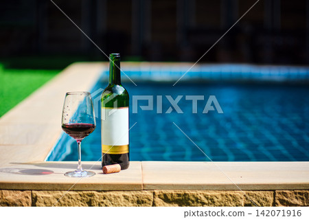 A bottle of red wine and a glass of wine are set on the edge of a clear blue pool, surrounded by a lush green lawn. The afternoon sun shines down, creating a relaxing and inviting atmosphere. A bottle of red wine and a glass of wine are set on the edge of a clear blue pool, surrounded by a lush green lawn. The afternoon sun shines down, creating a relaxing and inviting atmosphere. 142071916