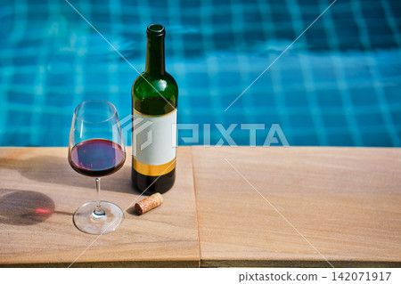 A bottle of red wine and a glass of wine are set on the edge of a clear blue pool, surrounded by a lush green lawn. The afternoon sun shines down, creating a relaxing and inviting atmosphere. A bottle of red wine and a glass of wine are set on the edge of a clear blue pool, surrounded by a lush green lawn. The afternoon sun shines down, creating a relaxing and inviting atmosphere. 142071917
