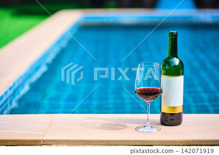 A bottle of red wine and a glass of wine are set on the edge of a clear blue pool, surrounded by a lush green lawn. The afternoon sun shines down, creating a relaxing and inviting atmosphere. A bottle of red wine and a glass of wine are set on the edge of a clear blue pool, surrounded by a lush green lawn. The afternoon sun shines down, creating a relaxing and inviting atmosphere. 142071919