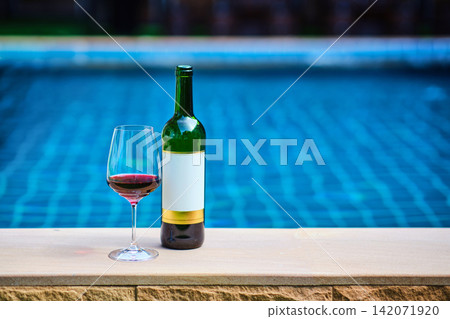 A bottle of red wine and a glass of wine are set on the edge of a clear blue pool, surrounded by a lush green lawn. The afternoon sun shines down, creating a relaxing and inviting atmosphere. A bottle of red wine and a glass of wine are set on the edge of a clear blue pool, surrounded by a lush green lawn. The afternoon sun shines down, creating a relaxing and inviting atmosphere. 142071920