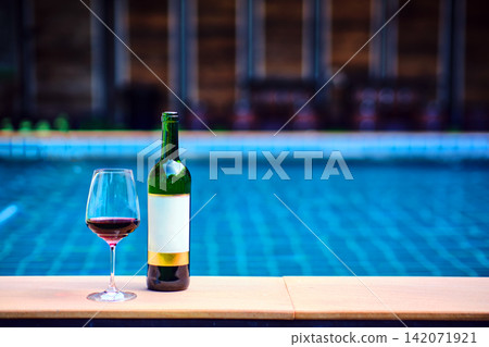 A bottle of red wine and a glass of wine are set on the edge of a clear blue pool, surrounded by a lush green lawn. The afternoon sun shines down, creating a relaxing and inviting atmosphere. 142071921