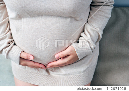 The elderly Asian woman with a pregnant belly sat on the gray sofa, gently cradling her growing baby inside. The elderly Asian woman with a pregnant belly sat on the gray sofa, gently cradling her growing baby inside. 142071926