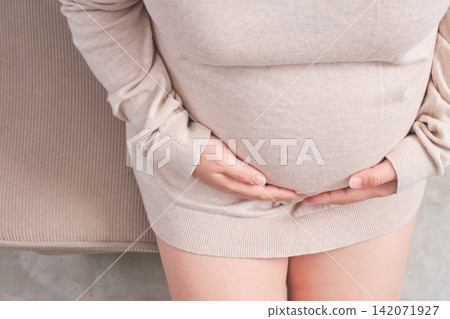 The elderly Asian woman with a pregnant belly sat on the gray sofa, gently cradling her growing baby inside. The elderly Asian woman with a pregnant belly sat on the gray sofa, gently cradling her growing baby inside. 142071927