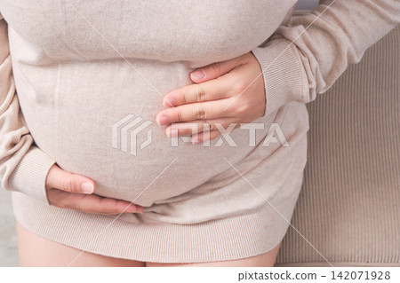 The elderly Asian woman with a pregnant belly sat on the gray sofa, gently cradling her growing baby inside. 142071928