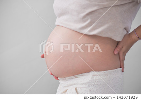 The mature Asian woman stood on the white backdrop, gracefully unveiling her large round belly, cradling it with both of her hands. 142071929