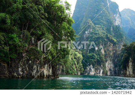 Rocky mountains, lush with trees, rise from the azure water, their cliffs catching the afternoon sun, set against a sky filled with clouds. Rocky mountains, lush with trees, rise from the azure water, their cliffs catching the afternoon sun, set against a sky filled with clouds. 142071942