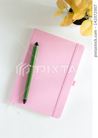 Pink notebook cover with green pen and yellow leaves plant on white desk Pink notebook cover with green pen and yellow leaves plant on white desk 142072007