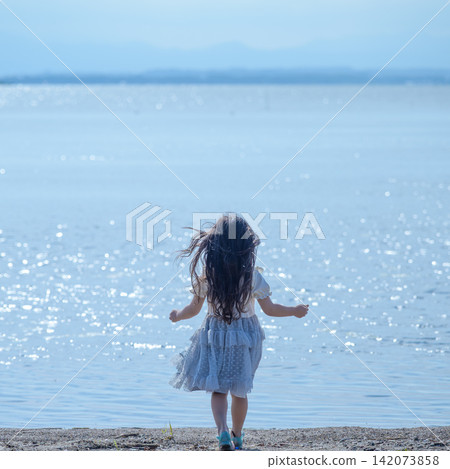 [Square] A girl in a white dress walks along a lakeside shimmering in the backlight | Her hair blowing in the wind, seen from behind. 142073858