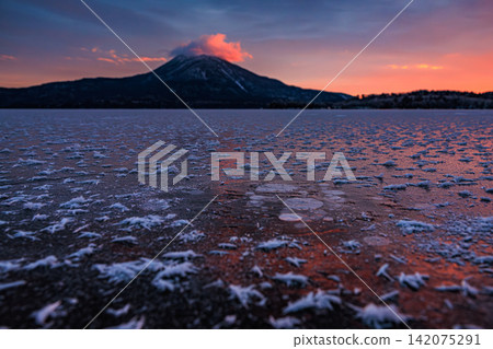 A winter morning scene woven from the crimson morning sky and ice bubbles and frost flowers trapped within the frozen Lake Akan. A winter morning scene woven from the crimson morning sky and ice bubbles and frost flowers trapped within the frozen Lake Akan. 142075291