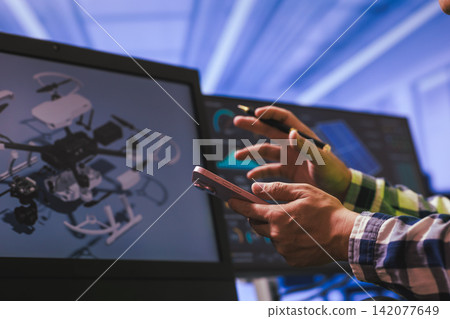 Hands using a smartphone near computer screens with a drone design on display, digital work, review, and modern technology in an office setting 142077649