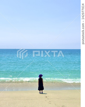 A girl's back is shown as she gazes out at the vast ocean at Wakimisaki Beach in Nagasaki City, against the backdrop of a summer blue sky and emerald green beach. A girl's back is shown as she gazes out at the vast ocean at Wakimisaki Beach in Nagasaki City, against the backdrop of a summer blue sky and emerald green beach. 142077814