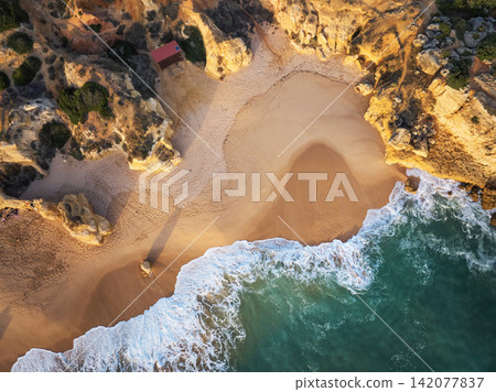 Drone top down view of a sandy cove, foamy surf, and golden cliffs at Praia de Sao Rafael beach. Drone top down view of a sandy cove, foamy surf, and golden cliffs at Praia de Sao Rafael beach. 142077837
