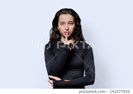 Attractive young fit woman holding finger on lips showing silence gesture against light background Attractive young fit woman holding finger on lips showing silence gesture against light background 142077850