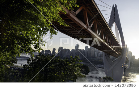 A dramatic low-angle shot reveals the bold orange-red truss and towering white pylon of the Qiansimen Bridge as it sweeps across the frame, set against the backdrop of Chongqing modern skyline. A dramatic low-angle shot reveals the bold orange-red truss and towering white pylon of the Qiansimen Bridge as it sweeps across the frame, set against the backdrop of Chongqing modern skyline. 142078090