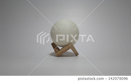 White spherical lamp resting on a small wooden tripod stand against a plain background, creating a minimalist interior object composition with modern design and soft neutral tones White spherical lamp resting on a small wooden tripod stand against a plain background, creating a minimalist interior object composition with modern design and soft neutral tones 142078096