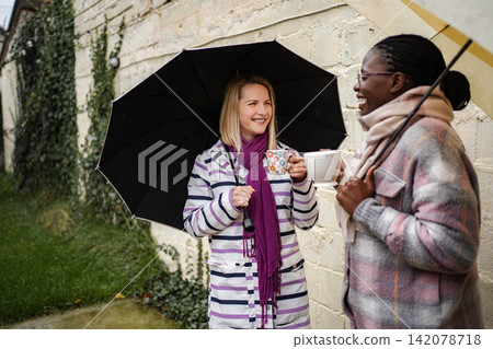 Two women enjoying coffee together under umbrellas during rain 142078718