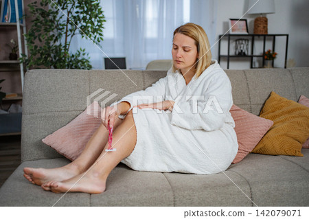 Woman in bathrobe shaving leg on living room couch Woman in bathrobe shaving leg on living room couch 142079071